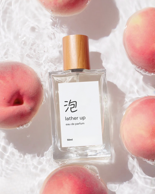 Perfume bottle labeled 'lather up' surrounded by peaches in shallow water.